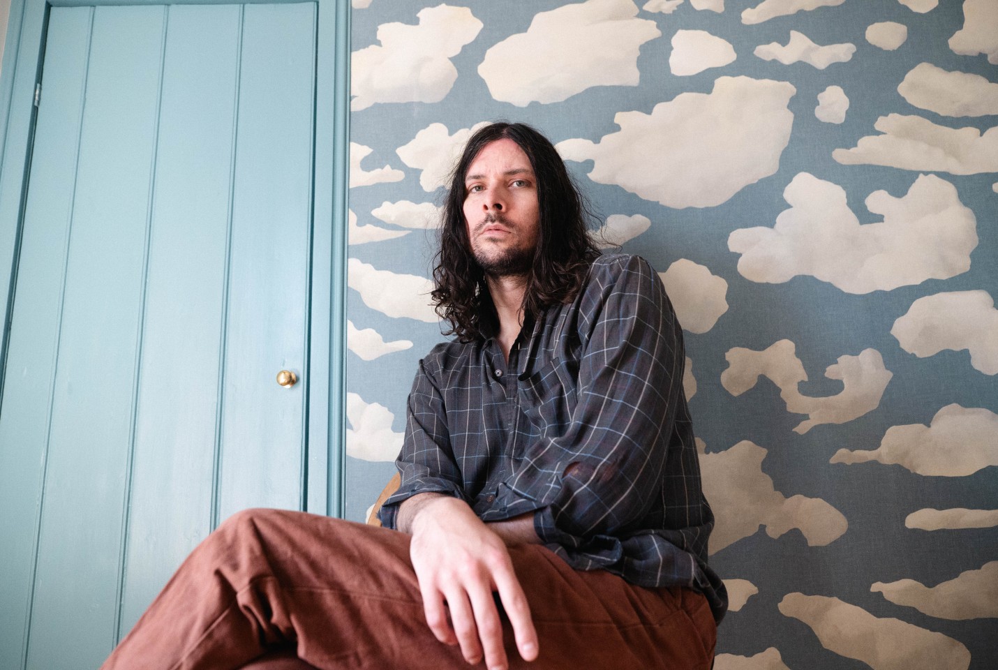 Man sitting against wall with long dark hair, wearing blue check shirt and brown trousers